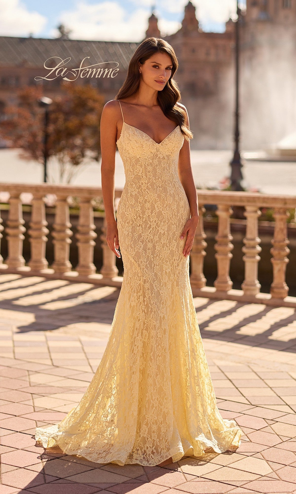 A woman in a pale yellow La Femme 33452 long prom dress with thin straps and a sheer back stands outdoors on a tiled patio, with a balustrade and fountain behind her.
